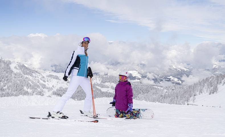 <p>Ein Kind auf einem Snowboard auf Knien vor einer Frau auf Skiern. Die Skipiste ist perfekt präpariert und die Landschaft rundherum tief verschneit. Der Himmel ist blau nur über dem Tal sind ein paar Wolken zu sehen. Dieses Bild entstand im Skigebiet Nassfeld in Kärnten, dem Skigebiet an der Grenze zu Italien, welches auch zu den "TOP 10 Skigebieten" Österreichs gehört.</p>