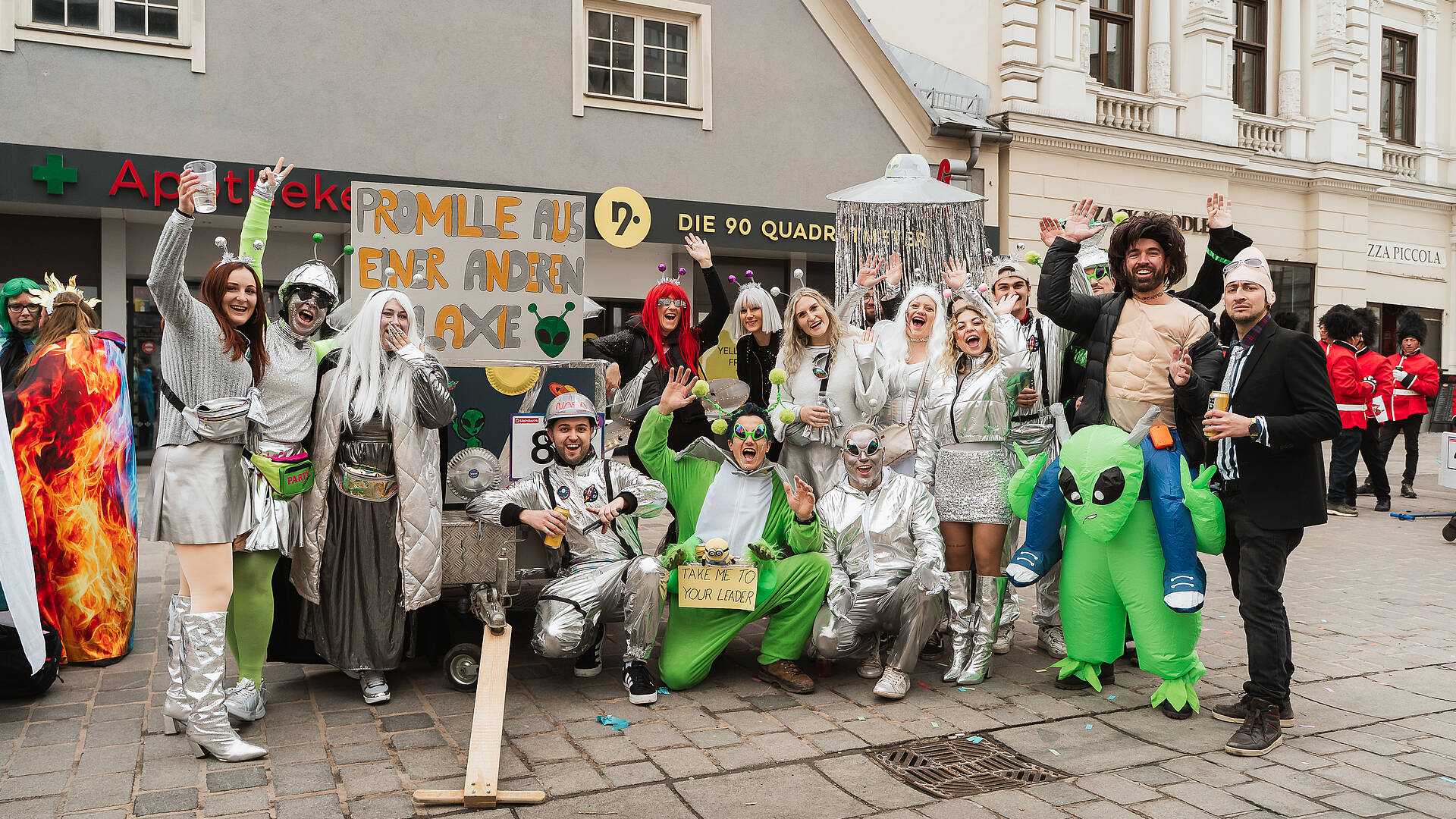 Gruppe von verkleideten Personen in Alien- und Raumanzugkostümen posiert lachend beim Villacher Fasching in der Innenstadt von Villach.