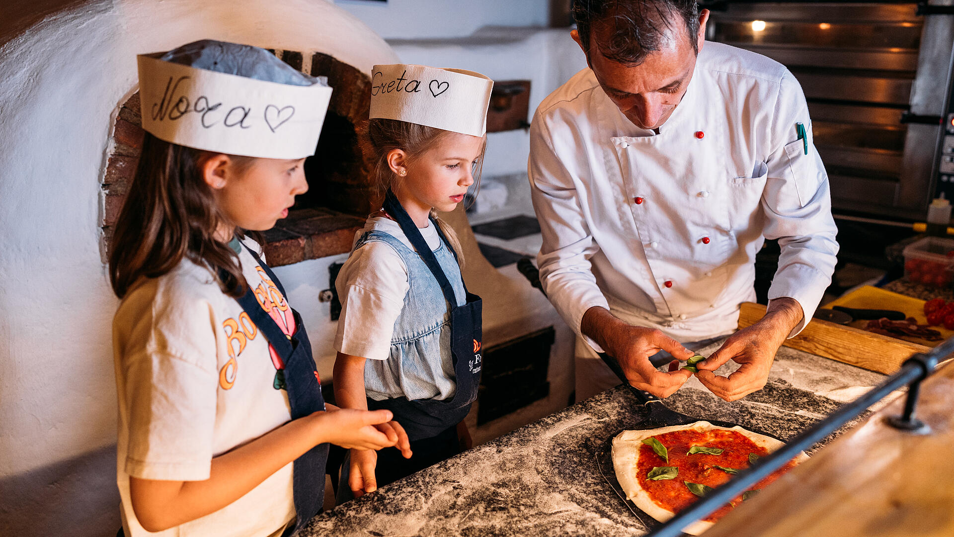Pizza backen wie die Profis – Kinderkurs im Trattlerhof Ein Koch zeigt zwei Mädchen, wie man eine Pizza mit Tomatensauce und Basilikum belegt. Die Kinder tragen Papierkochmützen mit ihren Namen und beobachten aufmerksam.