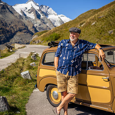 Ein beige‑brauner Oldtimer parkt vor einer eindrucksvollen Bergkulisse mit klar erkennbarer Gletscherregion. Perfekt für Themen wie Nostalgie, Alpenpanorama und Entdeckertouren in Kärnt