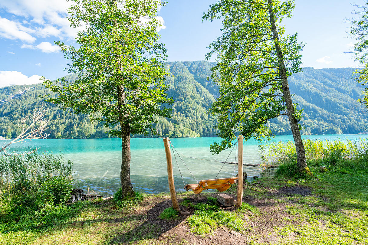 Holzschaukel zwischen zwei Bäumen direkt am Ufer des Weissensees mit Blick auf das klare Wasser und die umliegenden Berge. Holzschaukel zwischen zwei Bäumen direkt am Ufer des Weissensees mit Blick auf das klare Wasser und die umliegenden Berge.