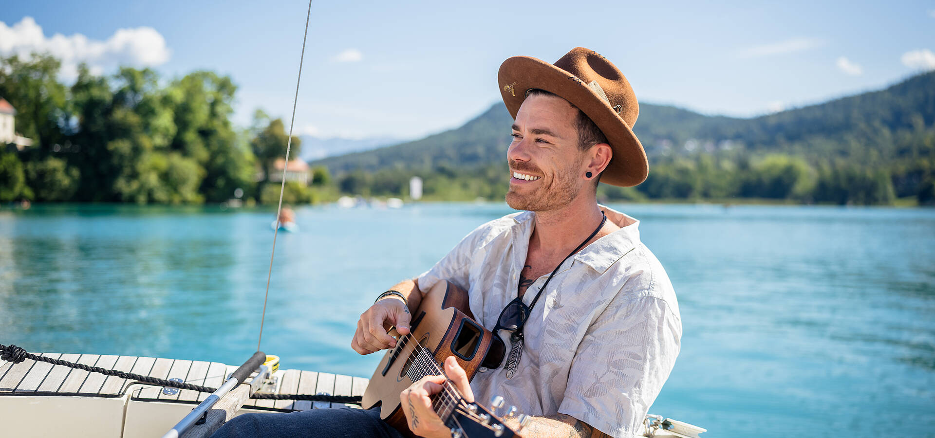 Eine Person sitzt auf einem Boot auf dem Wörthersee und spielt eine Akustikgitarre. Im Hintergrund spiegeln sich Wälder und Hügel im klaren Wasser. Die Szene verbindet Ruhe, Natur und kreative Leichtigkeit – ein typischer Kärntner Sommermoment.