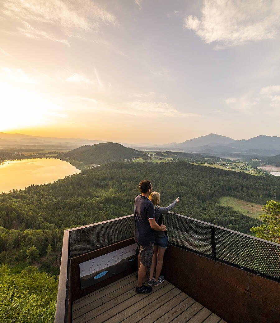 Zwei Personen stehen auf einer hölzernen Aussichtsplattform am Kitzelberg und blicken über eine ausgedehnte Seen‑ und Waldlandschaft, die im warmen Abendlicht leuchtet. Die Szene zeigt einen erhöhten Aussichtsort, der weite Panoramen, stille Natur und klare Sicht über Täler und Gewässer vereint.