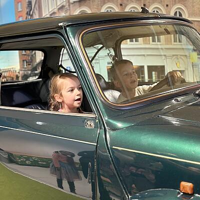 Zwei Kinder sitzen lachend in einem grünen Oldtimer im Indoor-Bereich von Minimundus Klagenfurt.