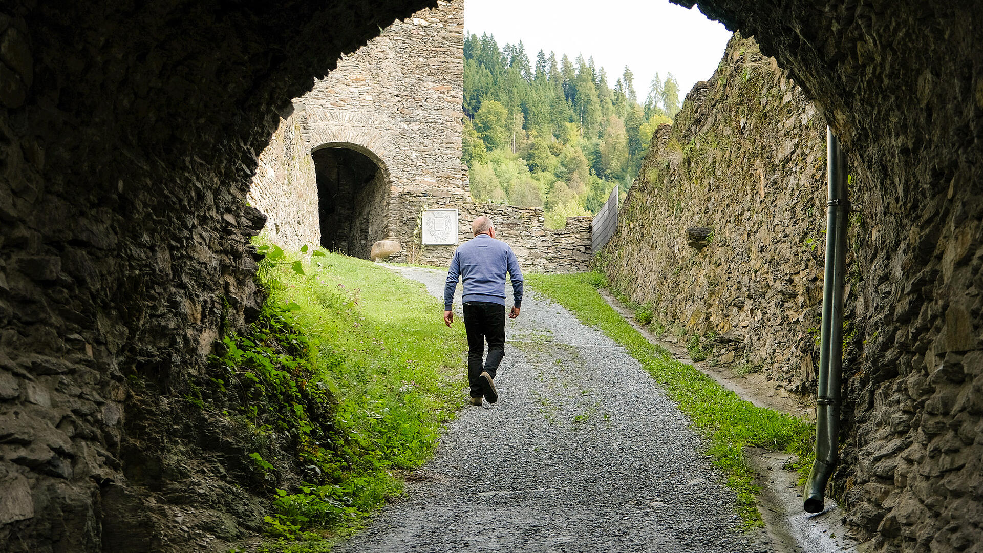 Franz Laßnig geht durch ein steinernes Tor der Burg Glanegg, flankiert von Burgmauern, im Hintergrund Waldhügel.