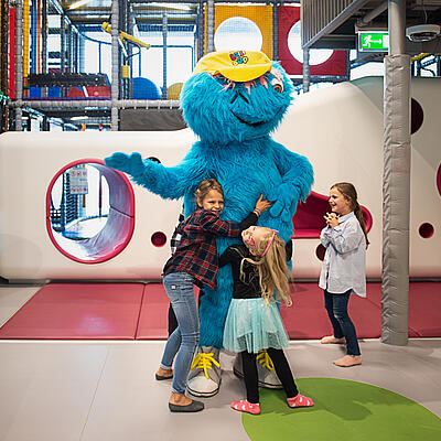 Drei Kinder spielen lachend mit einem blauen Plüschmaskottchen im bunten Indoor-Spielbereich von Planet Lollipop