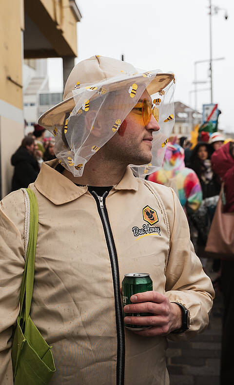 Mann mit Hut, Schleier und Bienenmotiv-Kostüm steht beim Villacher Fasching in der Innenstadt von Villach und hält eine Getränkedose in der Hand.