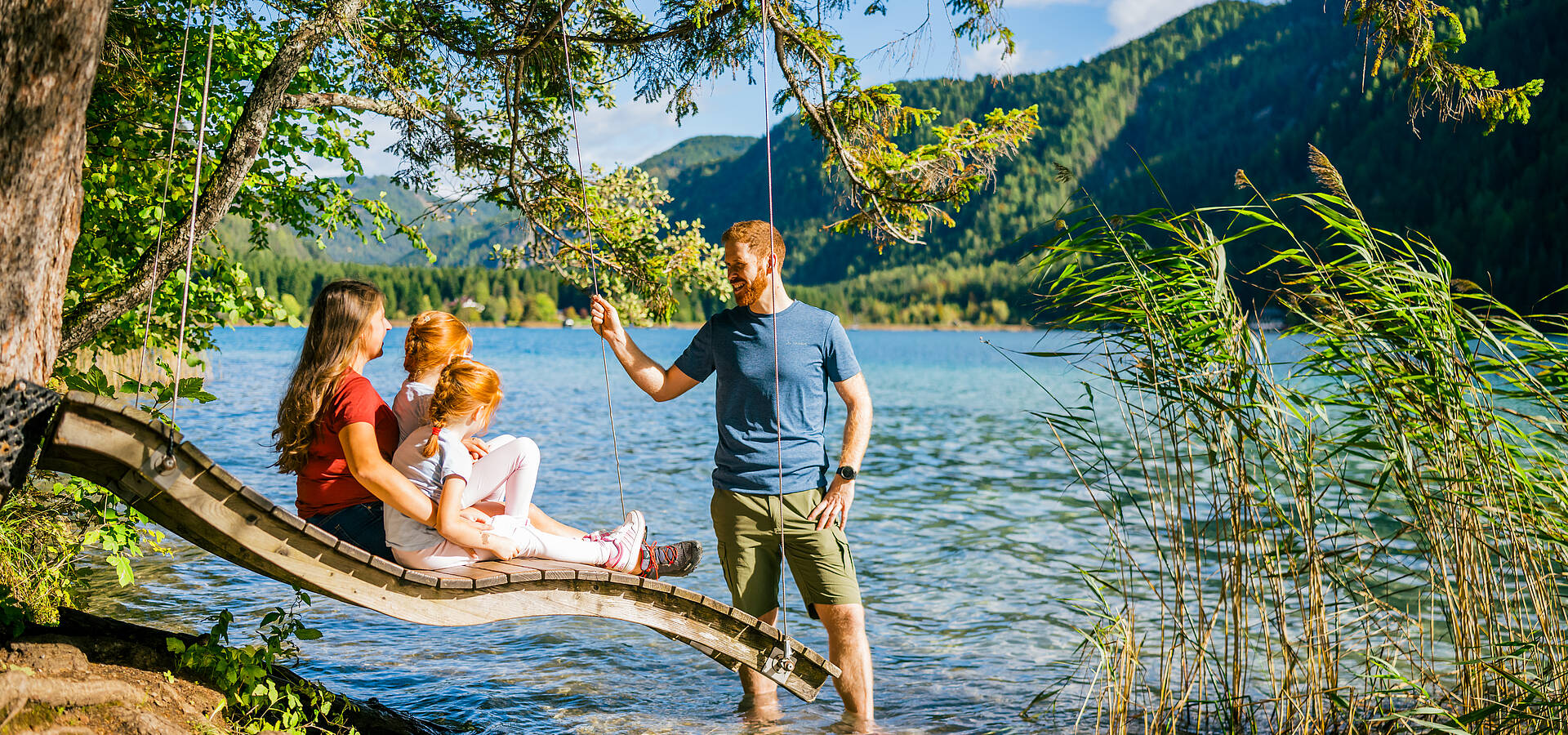 Eine Familie sitzt auf einer geschwungenen Holzplattform direkt am Ufer eines klaren Kärntner Sees, während eine weitere Person ihnen vom Ufer aus etwas zeigt. Umgeben von grünem Schilf, Waldhängen und Berglandschaft vermittelt das Bild Ruhe, Naturverbundenheit und die entspannte Atmosphäre der Kärntner Seenwelt.