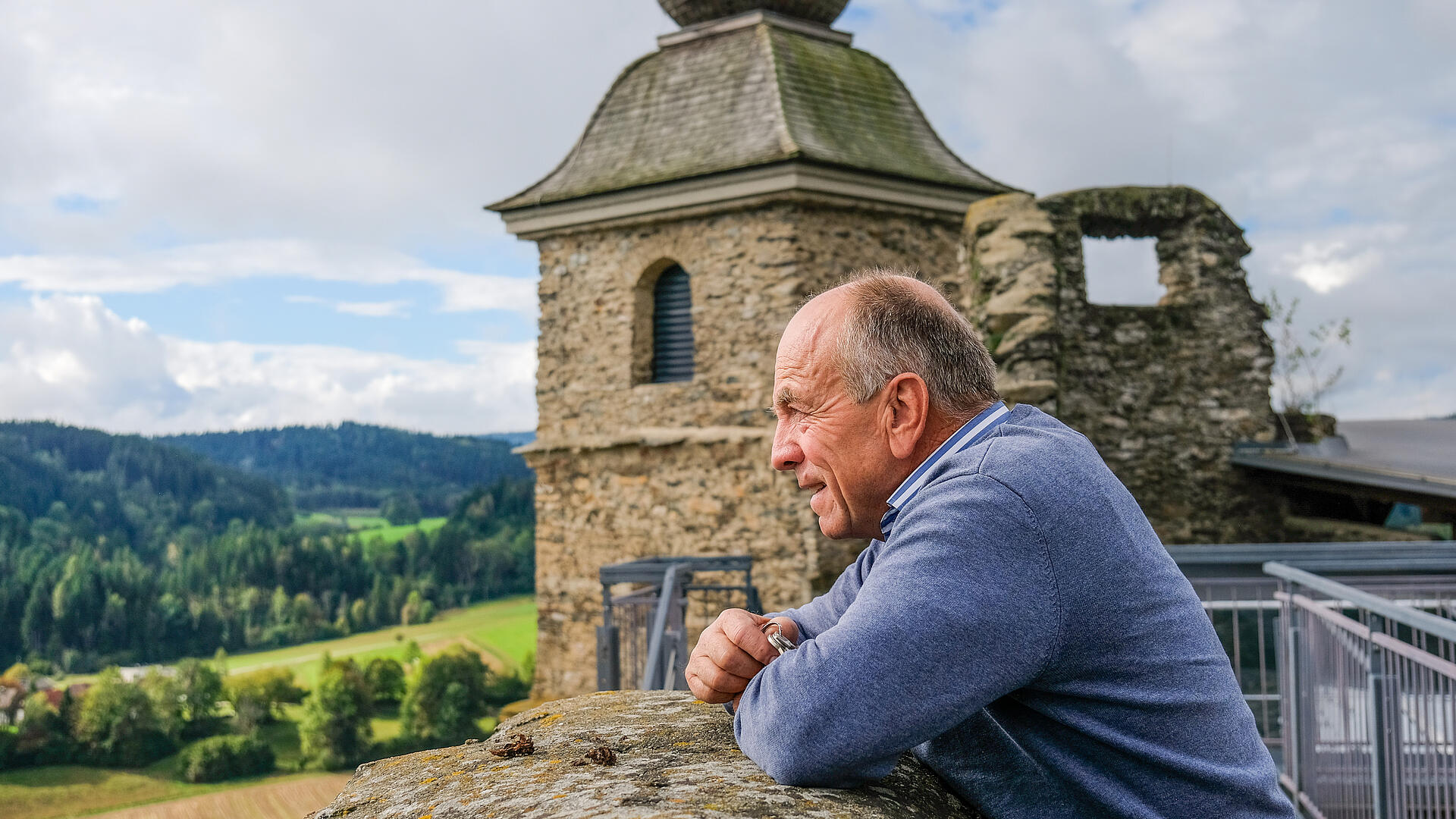 Franz Laßnig lehnt an der Mauer der Burg Glanegg und blickt über das grüne Tal und die umliegenden Wälder.