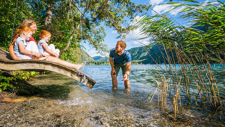 Eine Familie genießt den Blick über einen türkisfarbenen See, eingerahmt von Wäldern und Bergen. Die Szene zeigt natürliche Ruhe, gemeinsames Erleben und den besonderen Charme der Kärntner Seenlandschaft.