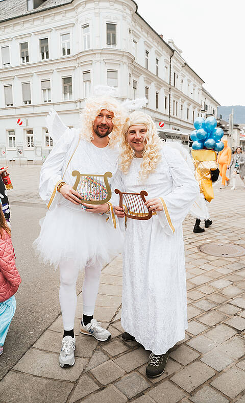 Zwei als Engel verkleidete Männer in weißen Gewändern und mit Harfen stehen beim Villacher Fasching in der Innenstadt von Villach.