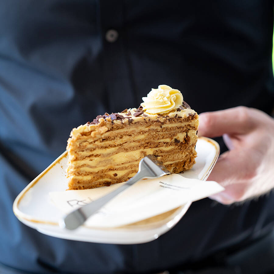 ine Person serviert ein Stück hausgemachter Torte auf einem Teller in einem Gasthof. Die Szene zeigt regionale Kulinarik, warme Atmosphäre und den einladenden Charakter eines traditionellen Kärntner Wirtshauses, in dem Genuss und Begegnung im Mittelpunkt stehen.