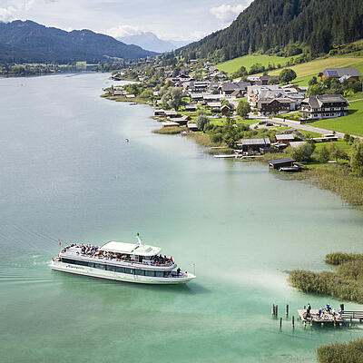 Ausflugsschiff fährt über das türkisfarbene Wasser des Weissensees, im Hintergrund das Ufer mit Häusern, Wiesen und Waldhängen.