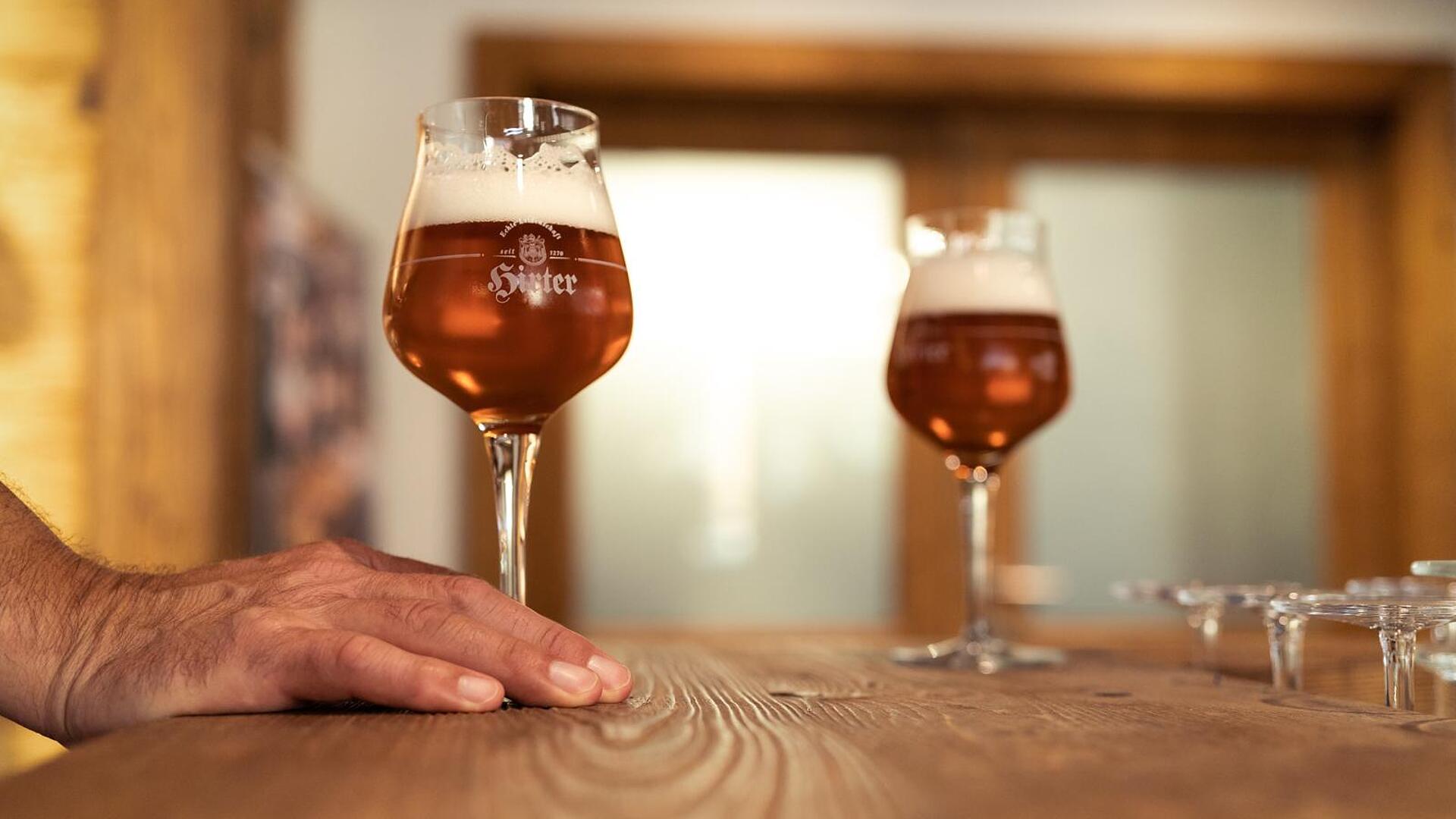 Privatbrauerei Hirt Zwei Gläser frisch gezapftes Bier in der Privatbrauerei Hirt, eines davon mit einer Hand auf dem Holztisch gehalten.