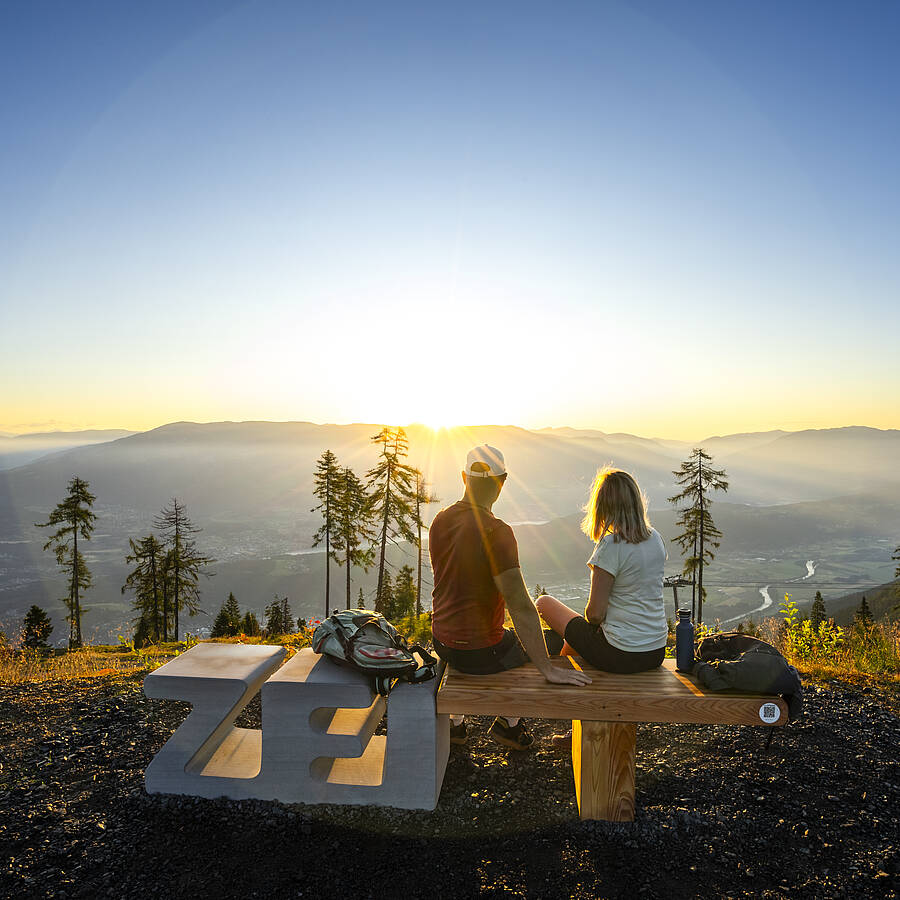 Zwei Personen sitzen auf einer Aussichtssitzbank am Goldeck und blicken auf das weitläufige Drautal, Wälder und Bergketten. Die tiefstehende Sonne beleuchtet die Landschaft und erzeugt eine klare, weite Panoramaansicht, die die Vielseitigkeit der Kärntner Berge zeigt.