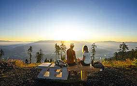 Zwei Personen sitzen auf einer Aussichtssitzbank am Goldeck und blicken auf das weitläufige Drautal, Wälder und Bergketten. Die tiefstehende Sonne beleuchtet die Landschaft und erzeugt eine klare, weite Panoramaansicht, die die Vielseitigkeit der Kärntner Berge zeigt.