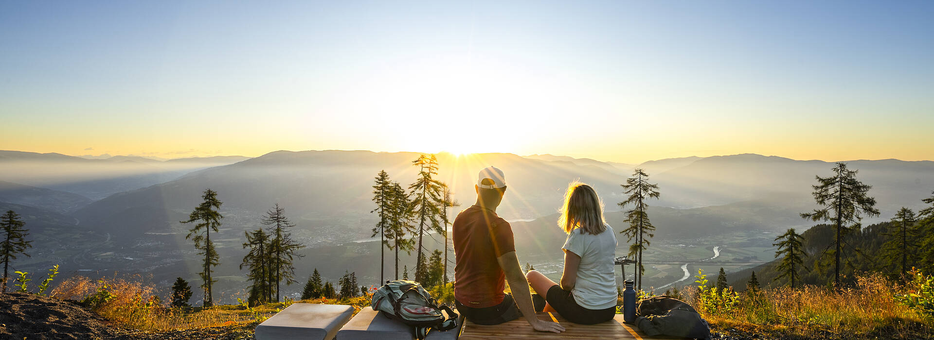 Zwei Personen sitzen auf einer Aussichtssitzbank am Goldeck und blicken auf das weitläufige Drautal, Wälder und Bergketten. Die tiefstehende Sonne beleuchtet die Landschaft und erzeugt eine klare, weite Panoramaansicht, die die Vielseitigkeit der Kärntner Berge zeigt.