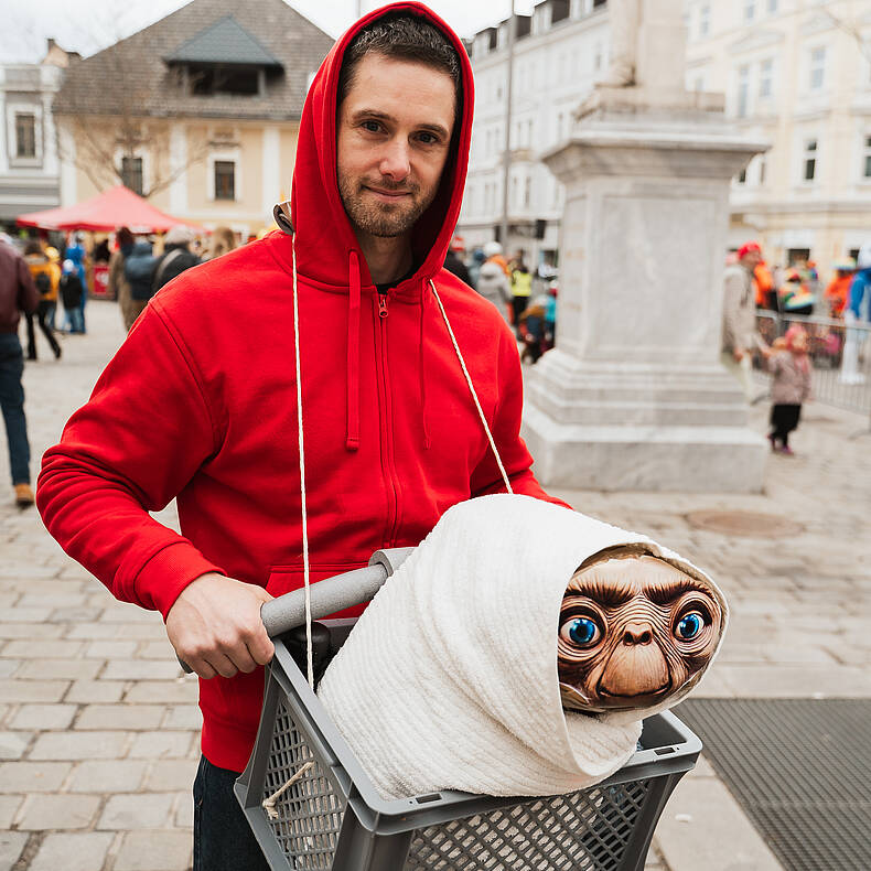 Mann in rotem Hoodie steht beim Villacher Fasching in der Innenstadt und hält eine Kiste mit einer als Filmfigur verkleideten Puppe.