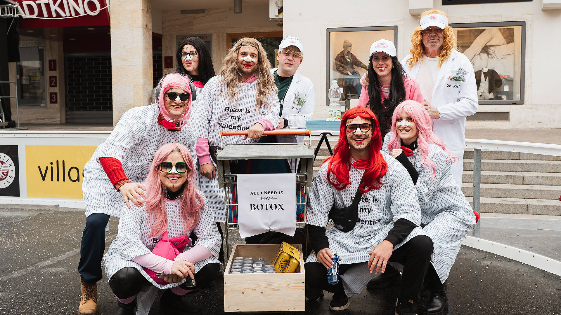Gruppe von verkleideten Personen in weißen Kitteln und bunten Perücken posiert beim Villacher Fasching vor einem Gebäude in der Innenstadt von Villach.