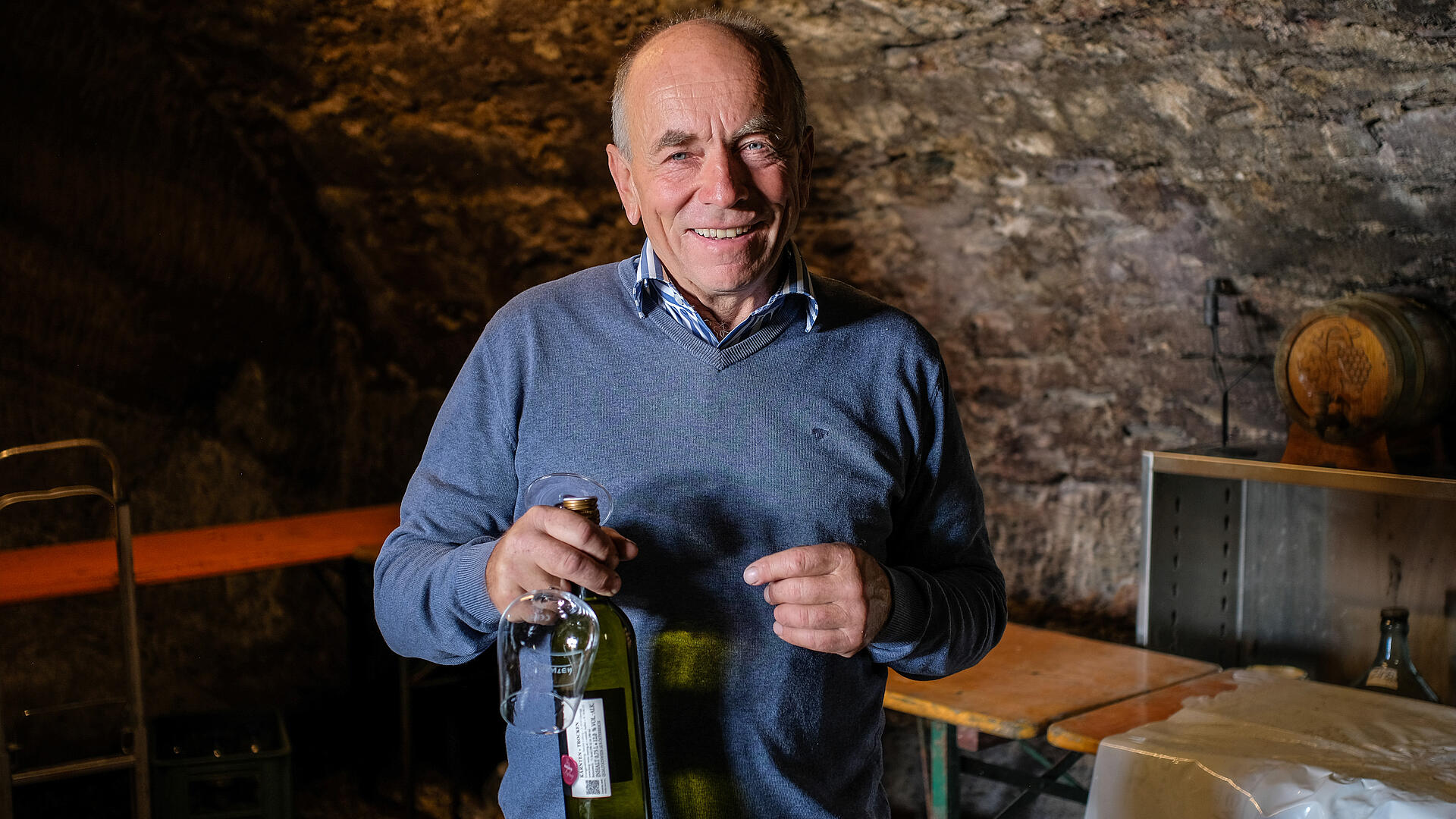 Franz Laßnig steht im Weinkeller der Buschenschank Laßnig und hält eine Flasche Burgwein mit Glas in der Hand.