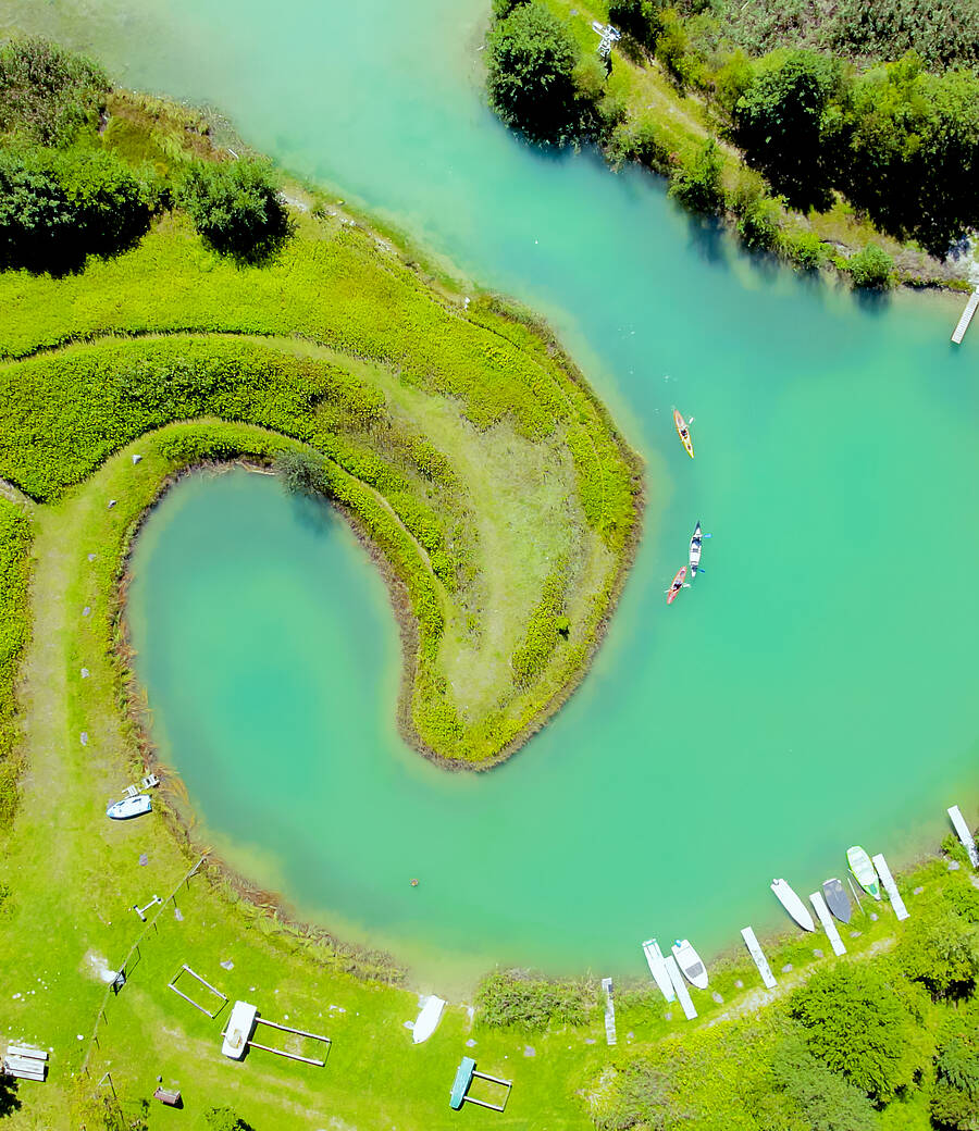 Ein versteckter Wasserarm in Ludmannsdorf windet sich hufeisenförmig durch eine grüne Uferlandschaft. Mehrere Boote und einzelne Personen auf dem Wasser zeigen die ruhige, abgeschiedene Atmosphäre des Platzes. Die Szene vermittelt Natur, Stille und einen Ort ideal zum Abschalten.