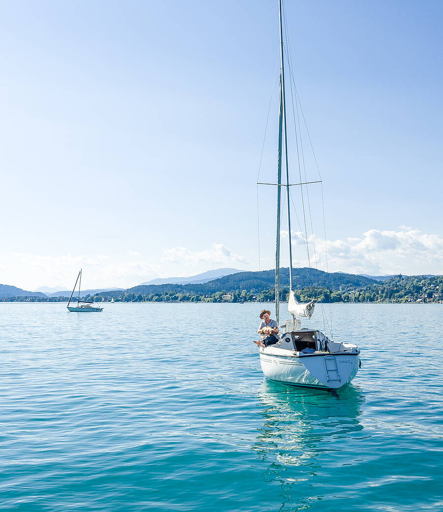 Auf einem Segelboot entsteht ein musikalischer Augenblick, eingerahmt vom ruhigen türkisfarbenen Wörthersee und der umliegenden Natur. Das Bild steht für Leichtigkeit, Naturverbundenheit und kreative Freiheit in Kärnten.
