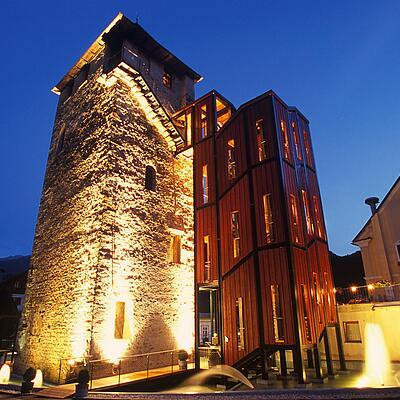 Historischer Mautturm in Winklern bei Nacht, stimmungsvoll beleuchtet, mit modernem Anbau und Wasserfontänen im Vordergrund.