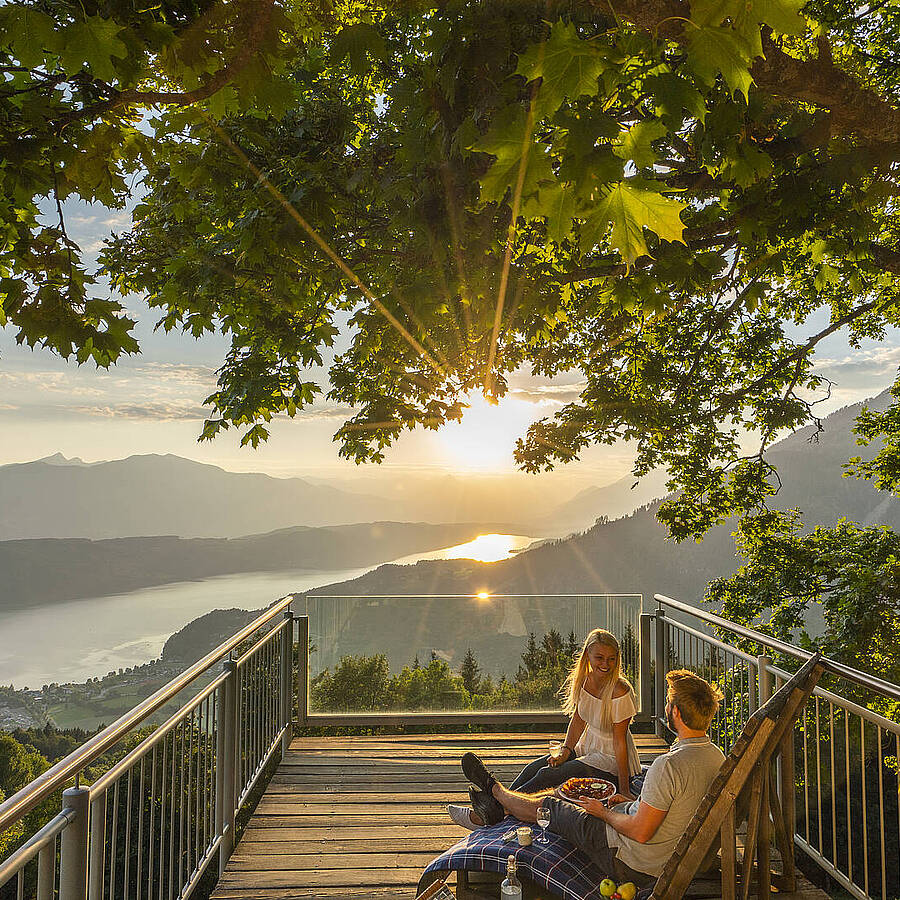 Zwei Personen sitzen auf einer Holzplattform unter einem schattenspendenden Baum und blicken über den Millstätter See, umgeben von Bergen, Tälern und warmem Abendlicht. Die Szene zeigt eine ruhige Aussichtsposition, die einen weiten Panoramablick und die natürliche Schönheit der Kärntner Landschaft hervorhebt.