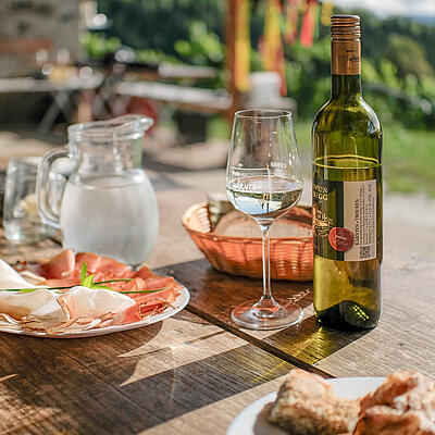 Tisch im Freien mit Burgwein Glanegg, Glas Wein, Kärntner Jause mit Speck und Brot, im Hintergrund grüne Landschaft.