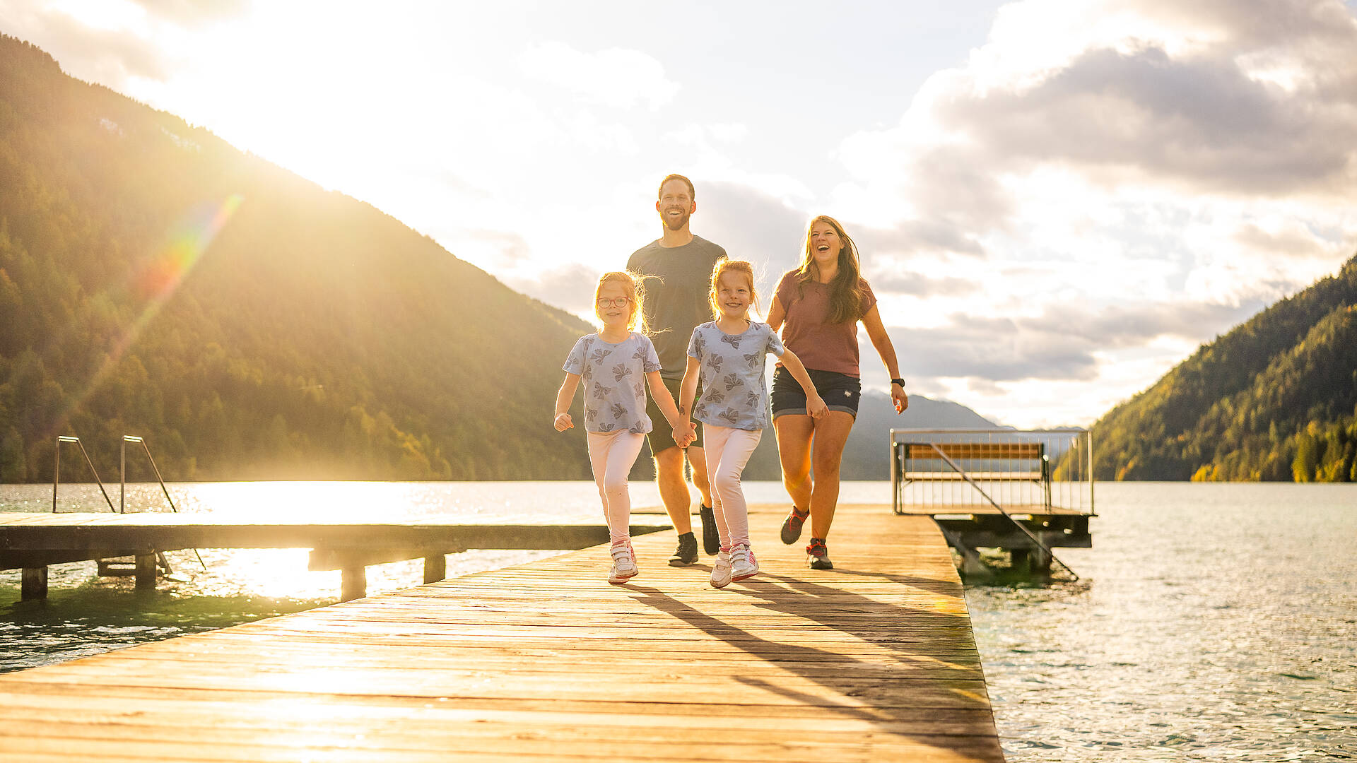 Eine Familie geht über einen Holzsteg am Weissensee, umgeben von klarem Wasser, bewaldeten Bergen und warmem Abendlicht. Die Szenerie zeigt die idyllische Naturkulisse des Weissensees und den besonderen Charakter der Kärntner Seenlandschaft.