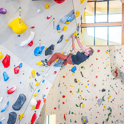 Mann klettert in der Indoor-Kletterhalle Villach an einer überhängenden Wand mit bunten Griffen, gesichert mit Klettergurt und Seil.