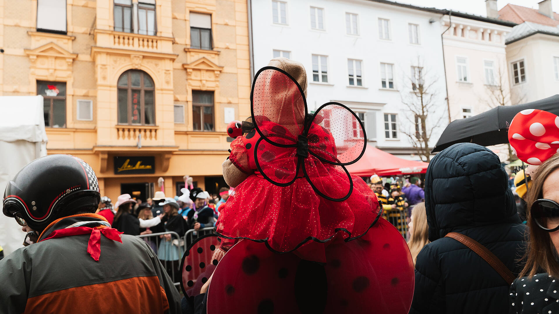 Kind im Marienkäferkostüm sitzt auf den Schultern eines verkleideten Elternteils beim Villacher Fasching in der Innenstadt von Villach.