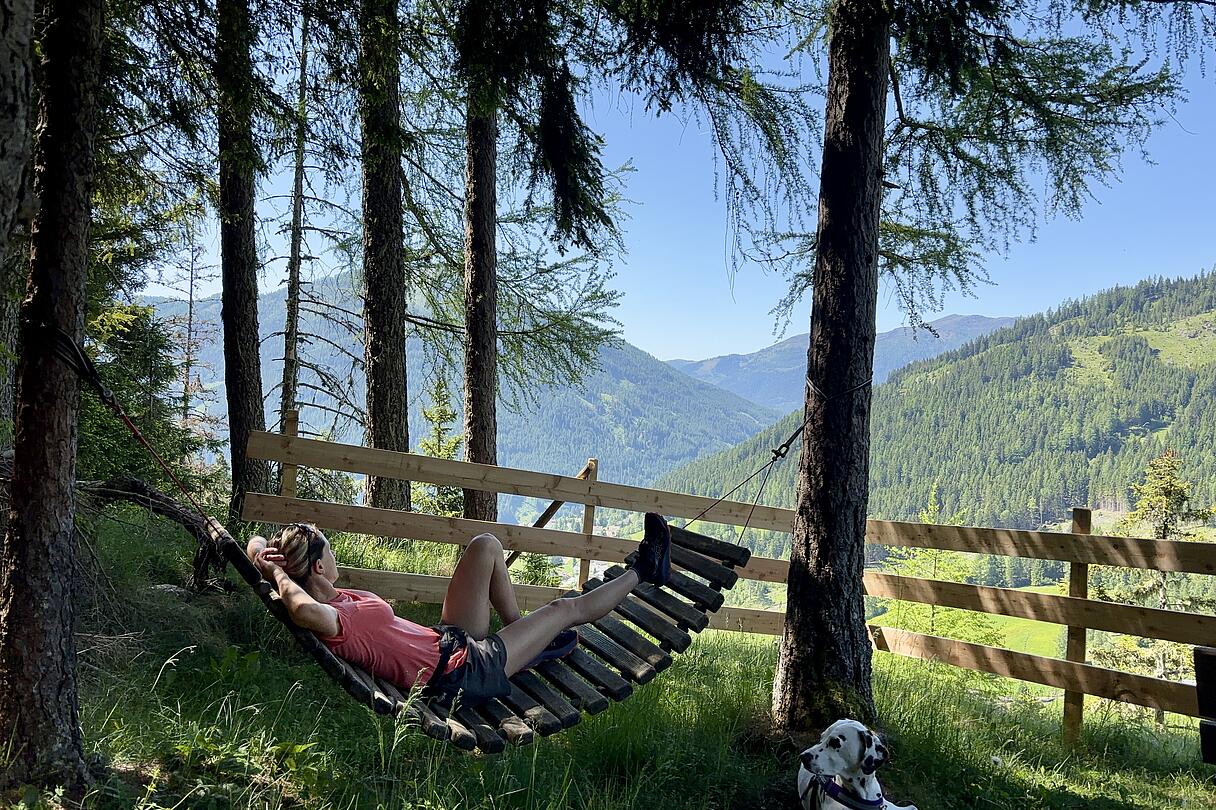 Weiße Wand am Hochsteig St. Oswald Frau entspannt in einer Hängeliege aus Holz zwischen Bäumen mit Blick auf die Berge, während ein Dalmatiner daneben liegt. Aufnahme an der Weißen Wand am Hochsteig St. Oswald.