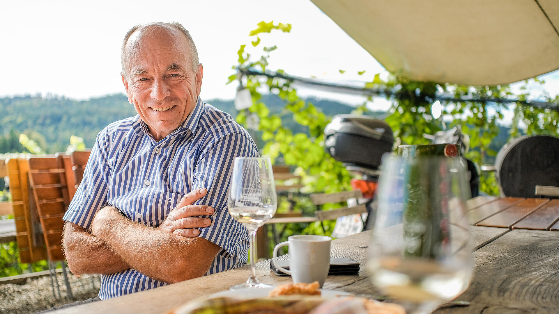 Franz Laßnig in der Buschenschank Laßnig im Freien Franz Laßnig sitzt lächelnd an einem Holztisch in der Buschenschank Laßnig, vor ihm ein Glas Wein und eine Kärntner Jause.