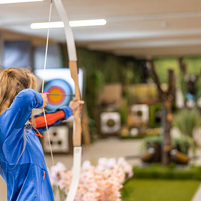 Junges Mädchen in blauer Sportkleidung zielt mit Pfeil und Bogen auf eine Zielscheibe in einer Indoor-Bogensportanlage