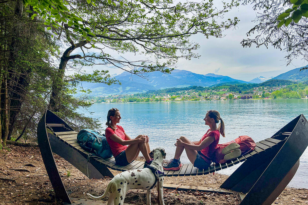 Slow Trail Millstätter See Südufer Zwei Frauen sitzen auf einer Holzliege am Südufer des Millstätter Sees, vor ihnen ein Dalmatiner, im Hintergrund Wasser und Berge.
