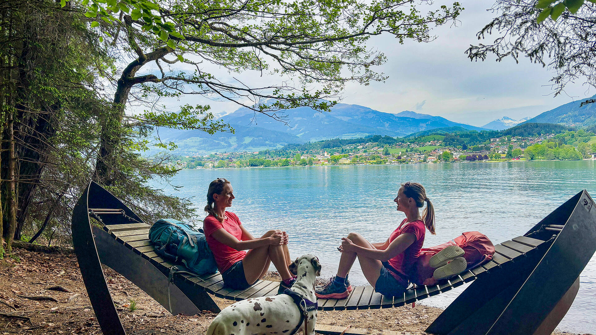 Zwei Frauen sitzen auf einer Holzliege am Südufer des Millstätter Sees, vor ihnen ein Dalmatiner, im Hintergrund Wasser und Berge.
