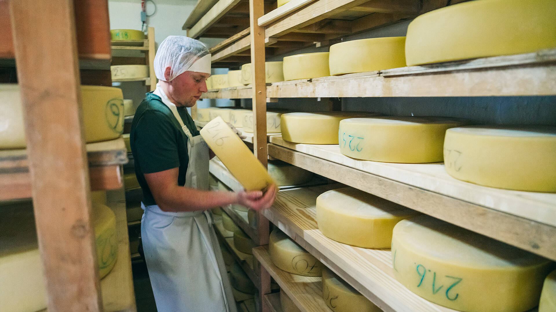 Der Senner bei der Arbeit, umgeben von Regalen voller reifer Käselaibe,