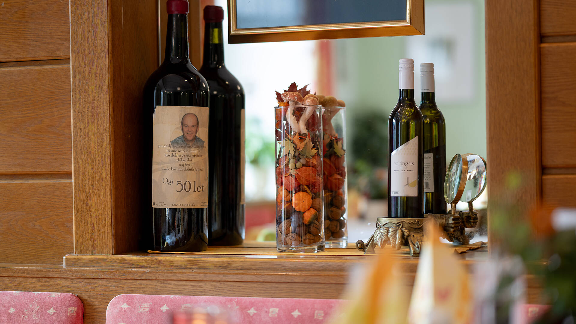 Zwei große Weinflaschen mit Etikett „Ogi 50 let“, herbstlich dekorierte Gläser mit Nüssen und Zierkürbissen, daneben zwei Weißweinflaschen mit dem Etikett „Ogris“, aufgestellt auf einem Holzregal im Gasthaus.