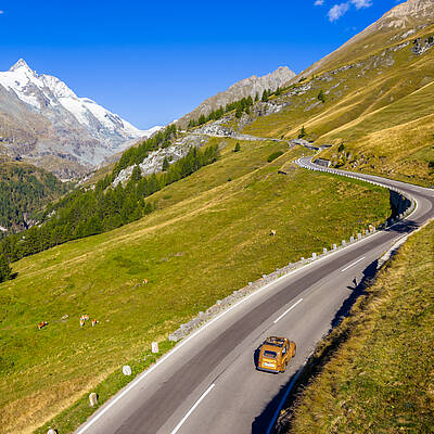 Ein Oldtimer fährt auf einer kurvenreichen Bergstraße oberhalb der Nockberge. Rechts und links erstrecken sich grüne Almflächen, während im Hintergrund ein markanter, schneebedeckter Gipfel sichtbar ist. Die Szene zeigt Weite, Höhenlage und die beeindruckende alpine Landschaft Kärntens.
