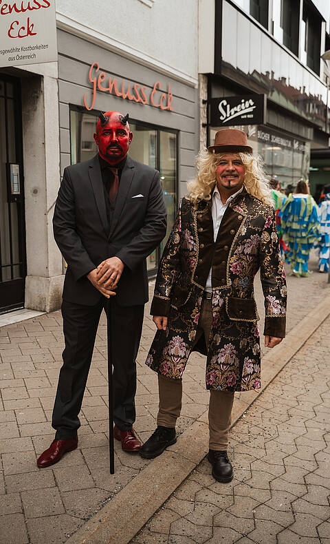 Zwei verkleidete Männer stehen beim Villacher Fasching in der Innenstadt von Villach, einer im Teufelskostüm mit rotem Gesicht, der andere in historisch anmutender Kleidung mit Hut.