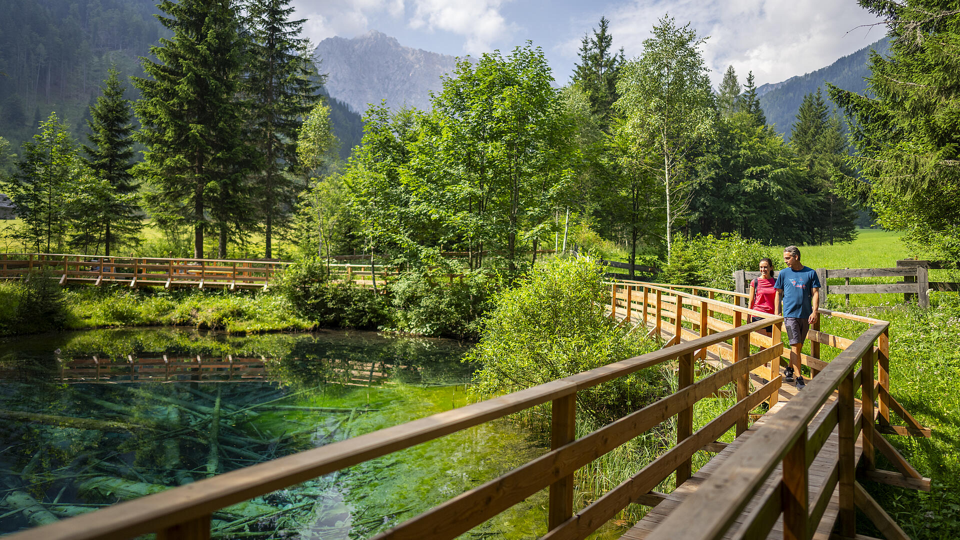 Meerauge im Bodental Zwei Wanderer spazieren auf einem Holzsteg entlang des türkisfarbenen Meerauges im Bodental, umgeben von grüner Natur, Bäumen und Bergkulisse.