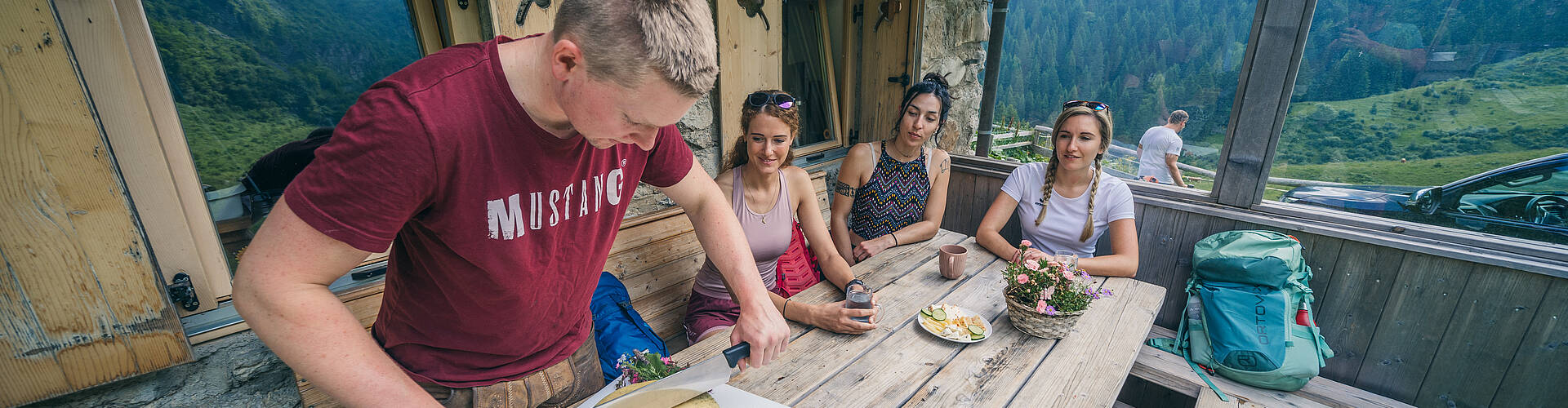Wanderer kehren in einer urigen Almhütte mit eigener Sennerei ein, wo ein großer, goldgelber Käselaib liebevoll angeschnitten wird und der Duft echter Almtradition die Stube erfüllt.