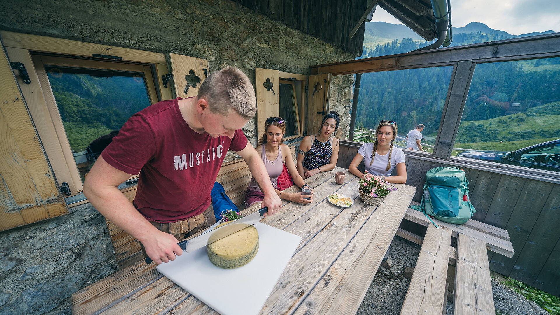 Wanderer kehren in einer urigen Almhütte mit eigener Sennerei ein, wo ein großer, goldgelber Käselaib liebevoll angeschnitten wird und der Duft echter Almtradition die Stube erfüllt.