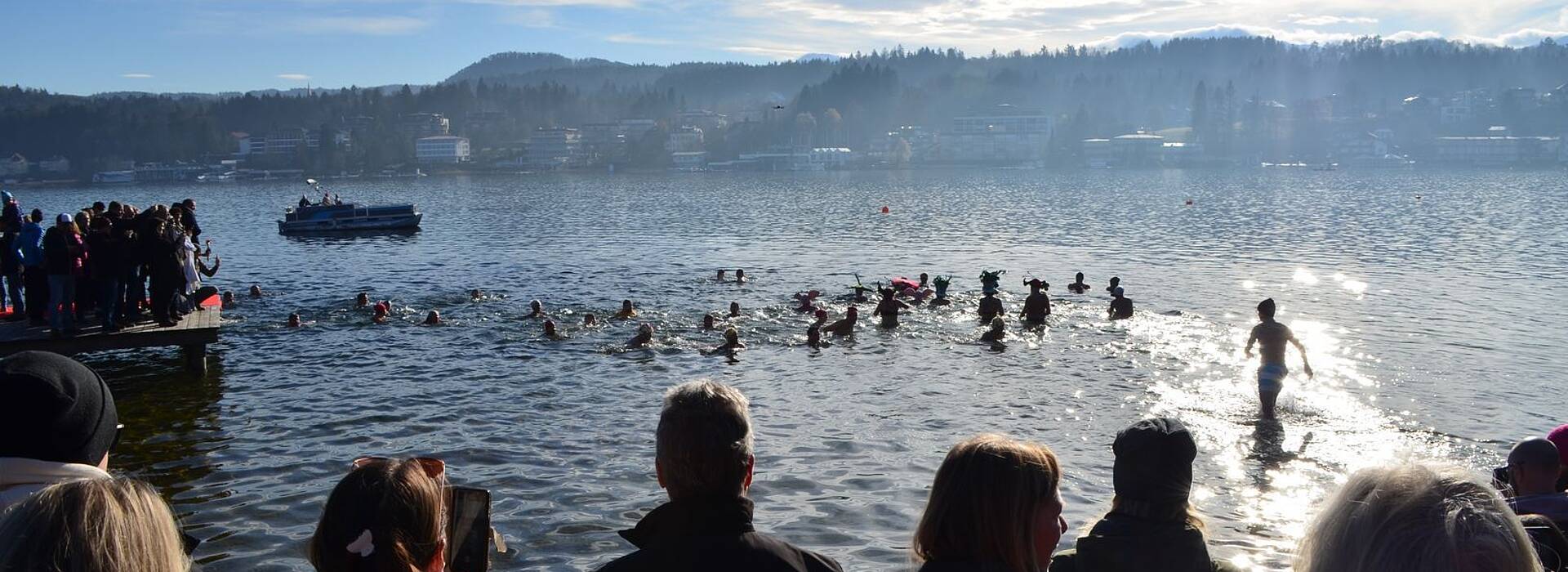 Zahlreiche Teilnehmer:innen, teils mit bunten Hauben, stürzen sich beim Neujahrsschwimmen am Wörthersee in den sonnig glitzernden Wörthersee. Dick eingepackte Zuschauer:innen verfolgen das Spektakel vom Ufer und Steg aus