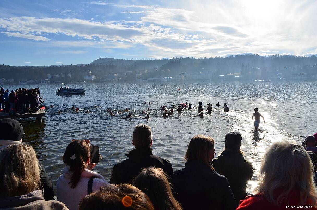 Zahlreiche Teilnehmer:innen, teils mit bunten Hauben, stürzen sich beim Neujahrsschwimmen am Wörthersee in den sonnig glitzernden Wörthersee. Dick eingepackte Zuschauer:innen verfolgen das Spektakel vom Ufer und Steg aus