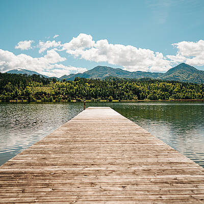 Ein Holzsteg führt in den Gösselsdorfer See, eingerahmt von klarem Wasser, bewaldeten Ufern und einer Bergkulisse unter blauem Himmel. Die Szene zeigt einen ruhigen Ort, ideal zum Durchatmen, Pausieren und den Blick über die Natur schweifen zu lassen.
